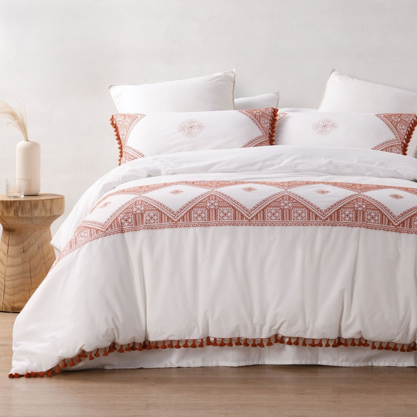 A white bedding set with a rust-orange geometric border pattern and matching tassels, styled on a bed with white pillows in a bright, minimalist bedroom setting.